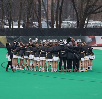 A group of athletes is gathered in a huddle on a sports field, wearing matching uniforms and gear. The ground is green, and there is signage with a brand logo in the background. Trees and parked cars can be seen behind a fence surrounding the field. A person on the left side is capturing the moment with a camera.