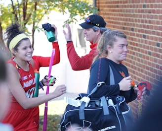 A group of athletes in sports attire appears to be in a joyful mood. One person is holding a field hockey stick and wearing a headband, while another carries a sports bag. They are gathered near a brick wall with trees in the background, sharing a light moment, likely after a sports event.