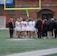 A group of athletes wearing sports uniforms huddles on a grassy field, likely preparing or strategizing for a game. The players are surrounded by additional team members or coaches wearing black jackets. A goalpost and net are visible in the background, along with a brick building.