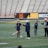 Several people are on a sports field with an empty bleacher in the background. One person is high-fiving a coach, while others stand nearby. They are wearing athletic clothing, hinting at a team sport environment. A sign with the text 'Muscle Milk' is visible in the background.