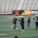 Several people are on a sports field with an empty bleacher in the background. One person is high-fiving a coach, while others stand nearby. They are wearing athletic clothing, hinting at a team sport environment. A sign with the text 'Muscle Milk' is visible in the background.